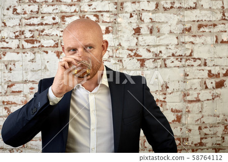 A man in a business suit on a background of a brick wall drinks a drink from a glass. The concept of A man in a business suit on a background of a brick wall drinks a drink from a glass. The concept of 58764112