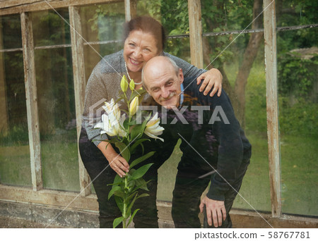 Old happy couple with flowers in a glass greenhouse near summer house 58767781