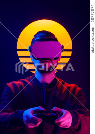 Man wearing virtual reality goggles playing with video game controller gamepad. VR head set videogame in 80's synth wave and retro wave futuristic aesthetics. 58772074
