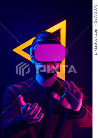Man wearing virtual reality goggles interacting with VR using hands motion control. VR head set videogame in 80's synth wave and retrowave glowing triangle futuristic aesthetics. Man wearing virtual reality goggles interacting with VR using hands motion control. VR head set videogame in 80's synth wave and retrowave glowing triangle futuristic aesthetics. 58772076