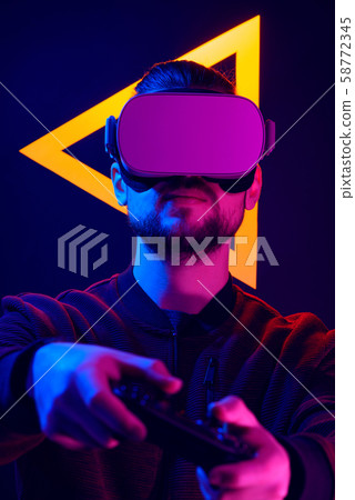 Man wearing virtual reality goggles interacting with VR using game pad wireless controller. VR headset videogame in 80's synth wave and retrowave glowing triangle futuristic aesthetics. 58772345