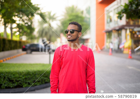 Happy young African bearded man with Afro hair thinking at the city streets Happy young African bearded man with Afro hair thinking at the city streets 58790819