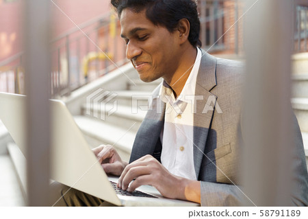 Young handsome Indian man is sitting with a laptop, smiling and typing Young handsome Indian man is sitting with a laptop, smiling and typing 58791189