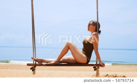 Woman in sunglasses and swimsuit is sitting on a swing on the beach Woman in sunglasses and swimsuit is sitting on a swing on the beach 58793714