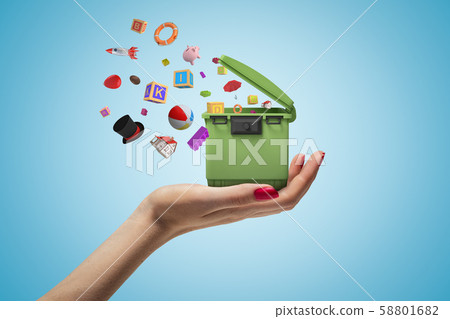Female hand holding green trash can with random objects falling out on blue background Female hand holding green trash can with random objects falling out on blue background 58801682