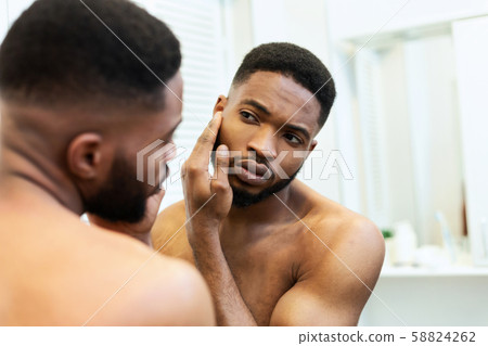 Young african american guy checking his face skin in mirror Young african american guy checking his face skin in mirror 58824262