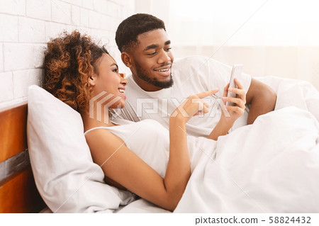 Cheerful african american couple networking on smartphone in bed Cheerful african american couple networking on smartphone in bed 58824432