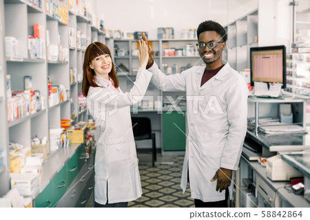Female and male pharmacists in pharmacy. African man and Caucasian woman giving five, working at Female and male pharmacists in pharmacy. African man and Caucasian woman giving five, working at 58842864