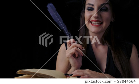 Beautiful sexy woman in a black witch costume and hat, conjures over a book with a pen, writes a pen Beautiful sexy woman in a black witch costume and hat, conjures over a book with a pen, writes a pen 58844564