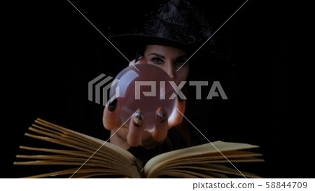 female in a black witch costume and hat, holds in her hands a transparent magic ball over a book and female in a black witch costume and hat, holds in her hands a transparent magic ball over a book and 58844709