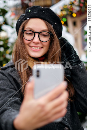 Beautiful young woman taking a selfie with christmas tree behind her with mobile phone camera - Beautiful young woman taking a selfie with christmas tree behind her with mobile phone camera - 58850933