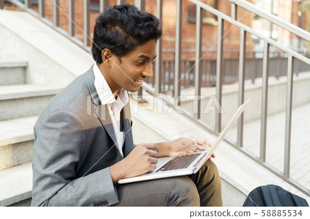 Young handsome Indian man is sitting with a laptop, smiling and typing Young handsome Indian man is sitting with a laptop, smiling and typing 58885534