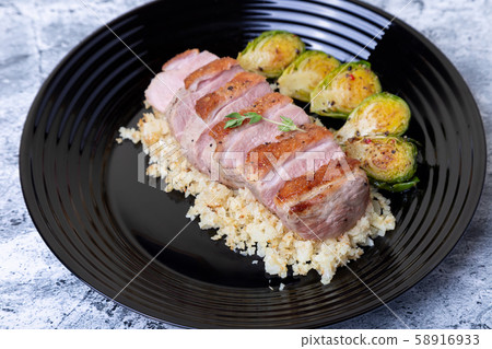 Grilled duck with fried Brussels sprouts, rice from cauliflower on a black plate. Close-up, selective focus. Grilled duck with fried Brussels sprouts, rice from cauliflower on a black plate. Close-up, selective focus. 58916933