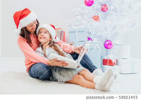 Pretty young mom reading an interesting book to her cute daughter near Christmas tree indoors. Pretty young mom reading an interesting book to her cute daughter near Christmas tree indoors. 58921455