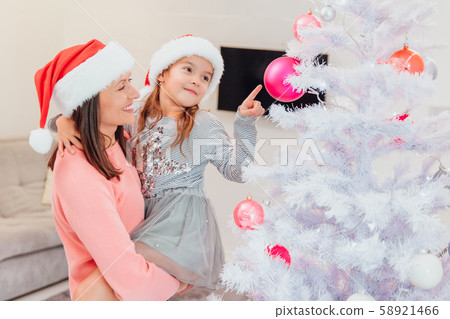 Mother holds her daughter, standing near christmas tree, cute girl is pointing at one of pink Mother holds her daughter, standing near christmas tree, cute girl is pointing at one of pink 58921466