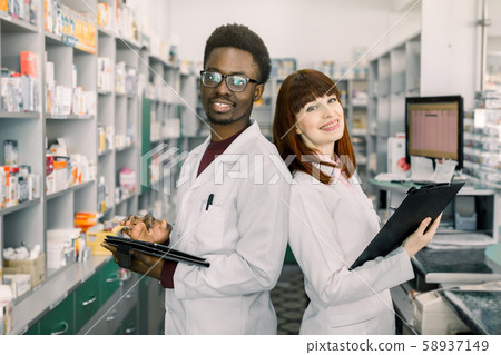 African man and Caucasian woman pharmacists are posing near table with cashbow in apothecary. African man and Caucasian woman pharmacists are posing near table with cashbow in apothecary. 58937149