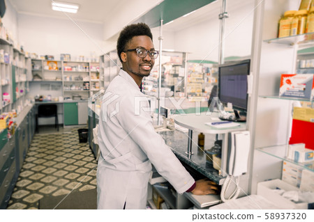 Portrait of young smiling African male pharmacist working with computer behind counter in pharmacy Portrait of young smiling African male pharmacist working with computer behind counter in pharmacy 58937320