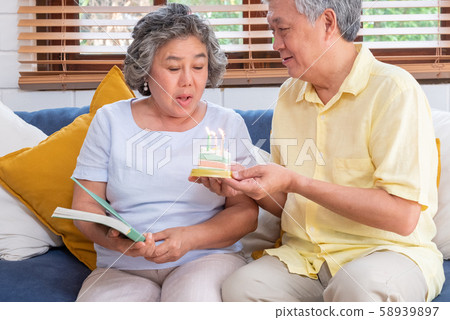 asian senior man surprise senior woman with birthday cake in living room at home asian senior man surprise senior woman with birthday cake in living room at home 58939897