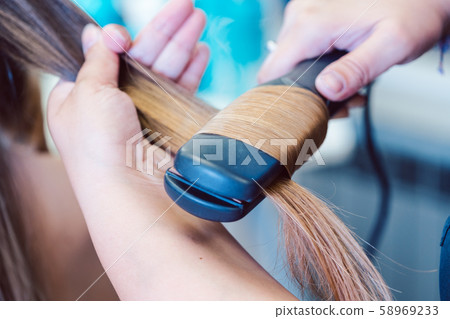Hairdresser using flat iron on hair of woman customer Hairdresser using flat iron on hair of woman customer 58969233