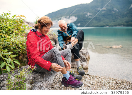 Senior pensioner couple hikers standing by lake in nature, taking shoes off. 58979366