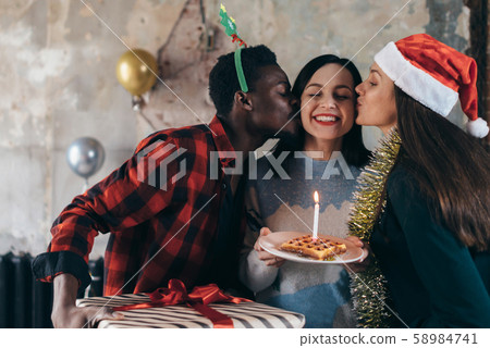 Woman holding birthday cake preparing to blow candle, friends around kissing cheeks. 58984741