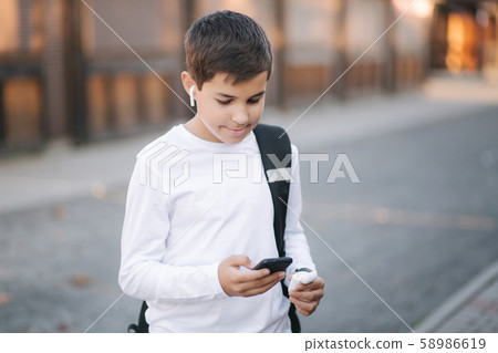 Teenage boy in white sweatshirt and with black backpuck use phone and wireless headphones and print Teenage boy in white sweatshirt and with black backpuck use phone and wireless headphones and print 58986619