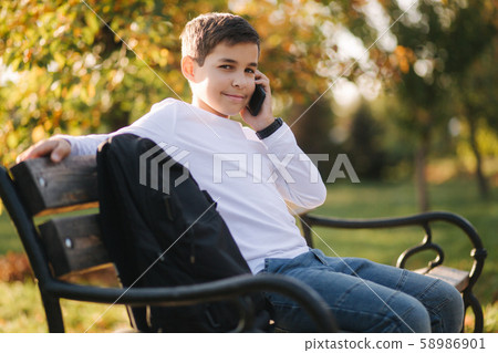 Young boy in white sweatshirt with black backpack sitting on the bench in the park and speak with 58986901