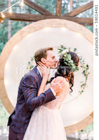 The newlywed stylish couple is hugging and kissing near the wedding arch in the restaurant in rustic 59014491