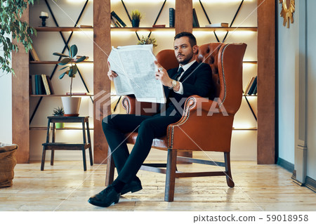 Confident young businessman reading newspaper and latest news while sitting in armchair Confident young businessman reading newspaper and latest news while sitting in armchair 59018958