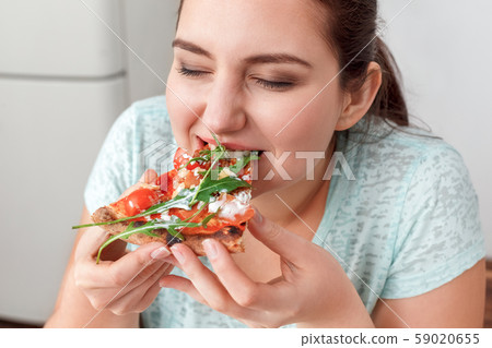 Binge Eating. Chubby girl sitting at kitchen table eating pizza closed eyes delightful close-up Binge Eating. Chubby girl sitting at kitchen table eating pizza closed eyes delightful close-up 59020655