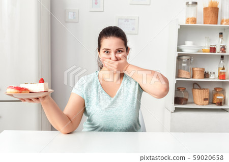 Overeating Problem. Chubby girl sitting at kitchen with plate of desserts stopping herself Overeating Problem. Chubby girl sitting at kitchen with plate of desserts stopping herself 59020658