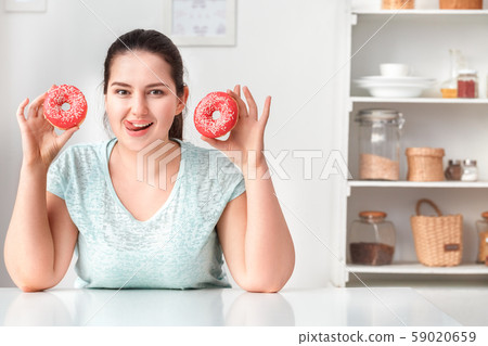 Junk Food. Chubby girl sitting at kitchen holding doughnuts showing tongue playful to camera Junk Food. Chubby girl sitting at kitchen holding doughnuts showing tongue playful to camera 59020659