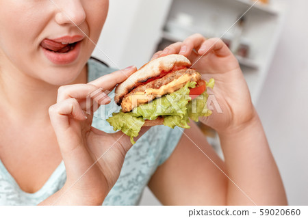 Junk Food. Chubby girl sitting at kitchen eating hamburger smiling playful close-up side view Junk Food. Chubby girl sitting at kitchen eating hamburger smiling playful close-up side view 59020660
