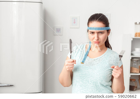 Be on Diet. Chubby girl with tape measure around head standing in kitchen with fork and knife Be on Diet. Chubby girl with tape measure around head standing in kitchen with fork and knife 59020662