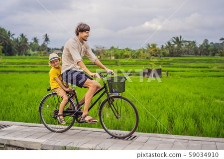 Father and son ride a bicycle on a rice field in Ubud, Bali. Travel to Bali with kids concept Father and son ride a bicycle on a rice field in Ubud, Bali. Travel to Bali with kids concept 59034910