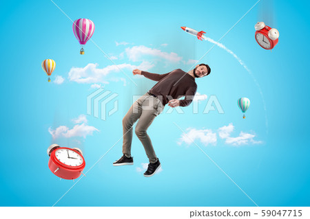 Young man and two alarm clocks floating in blue sky with rocket and hot air balloons in background. 59047715