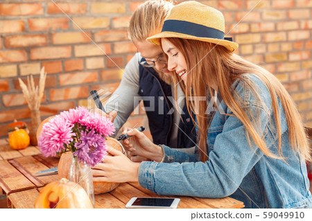 Halloween Preparaton Concept. Young couple sitting at table at porch making jack-o'-lantern drawing Halloween Preparaton Concept. Young couple sitting at table at porch making jack-o'-lantern drawing 59049910