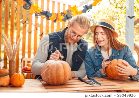 Halloween Preparaton Concept. Young couple sitting at table at porch making jack-o'-lantern man Halloween Preparaton Concept. Young couple sitting at table at porch making jack-o'-lantern man 59049926