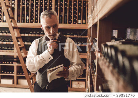 Sommelier Concept. Senior man standing with notebook checking wine collection pensive Sommelier Concept. Senior man standing with notebook checking wine collection pensive 59051770