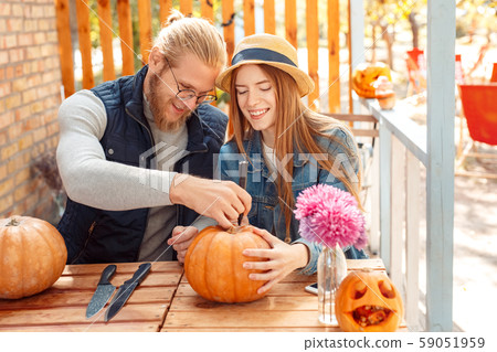 Halloween Preparaton Concept. Young couple sitting at table at porch making jack-o'-lantern carving Halloween Preparaton Concept. Young couple sitting at table at porch making jack-o'-lantern carving 59051959