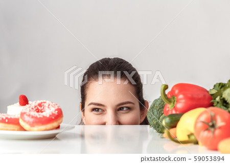 Body Care. Chubby girl standing hiding under kitchen table with vegetables looking at desserts 59053894