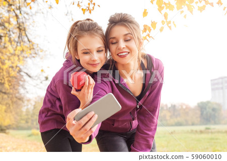 Outdoors leisure. Sisters standing hugging in the autumn park taking selfie on smartphone with apple 59060010