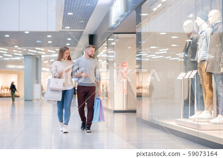 Young couple discussing stuff in shop window while passing by one of departments Young couple discussing stuff in shop window while passing by one of departments 59073164