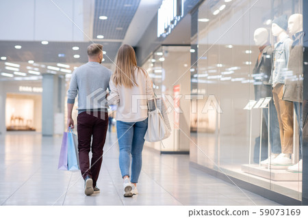 Rear view of young casual couple moving along shop window in the mall 59073169