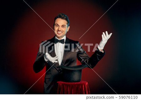 Professional Occupation. Magician in suit and gloves standing isolated on wall making trick with hat Professional Occupation. Magician in suit and gloves standing isolated on wall making trick with hat 59076110