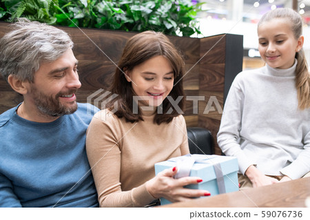 Young mother looking at giftbox in her hands surrounded by husband and daughter Young mother looking at giftbox in her hands surrounded by husband and daughter 59076736