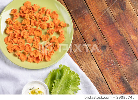 Round green plate with homemade carrot chips on, cup with sour cream sauce, green salad on white napkin on rustic wooden background. Natural healthy vegetable snack. Alternative to ready-made chips. Round green plate with homemade carrot chips on, cup with sour cream sauce, green salad on white napkin on rustic wooden background. Natural healthy vegetable snack. Alternative to ready-made chips. 59083743