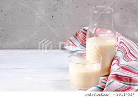 Close-up glass and bottle with lactose free oat milk and bright striped kitchen towel on white wooden tableon grey concrete wall background. Best dairy free substitutes of milk concept. Selective focu Close-up glass and bottle with lactose free oat milk and bright striped kitchen towel on white wooden tableon grey concrete wall background. Best dairy free substitutes of milk concept. Selective focu 59114534