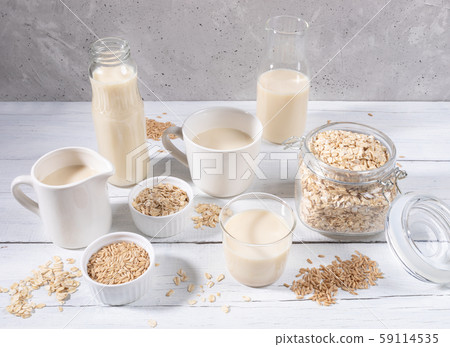 Set of different containers with healthy dairy free oat milk, oat seeds and flakes on white wooden table on grey concrete background. Lactose free substitutes of milk concept. 59114535