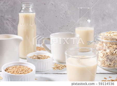 Close-up set of different glass containers with healthy dairy free oat milk, oat seeds and flakes on white painted wooden table on grey concrete background. Lactose free substitutes of milk concept. Close-up set of different glass containers with healthy dairy free oat milk, oat seeds and flakes on white painted wooden table on grey concrete background. Lactose free substitutes of milk concept. 59114536